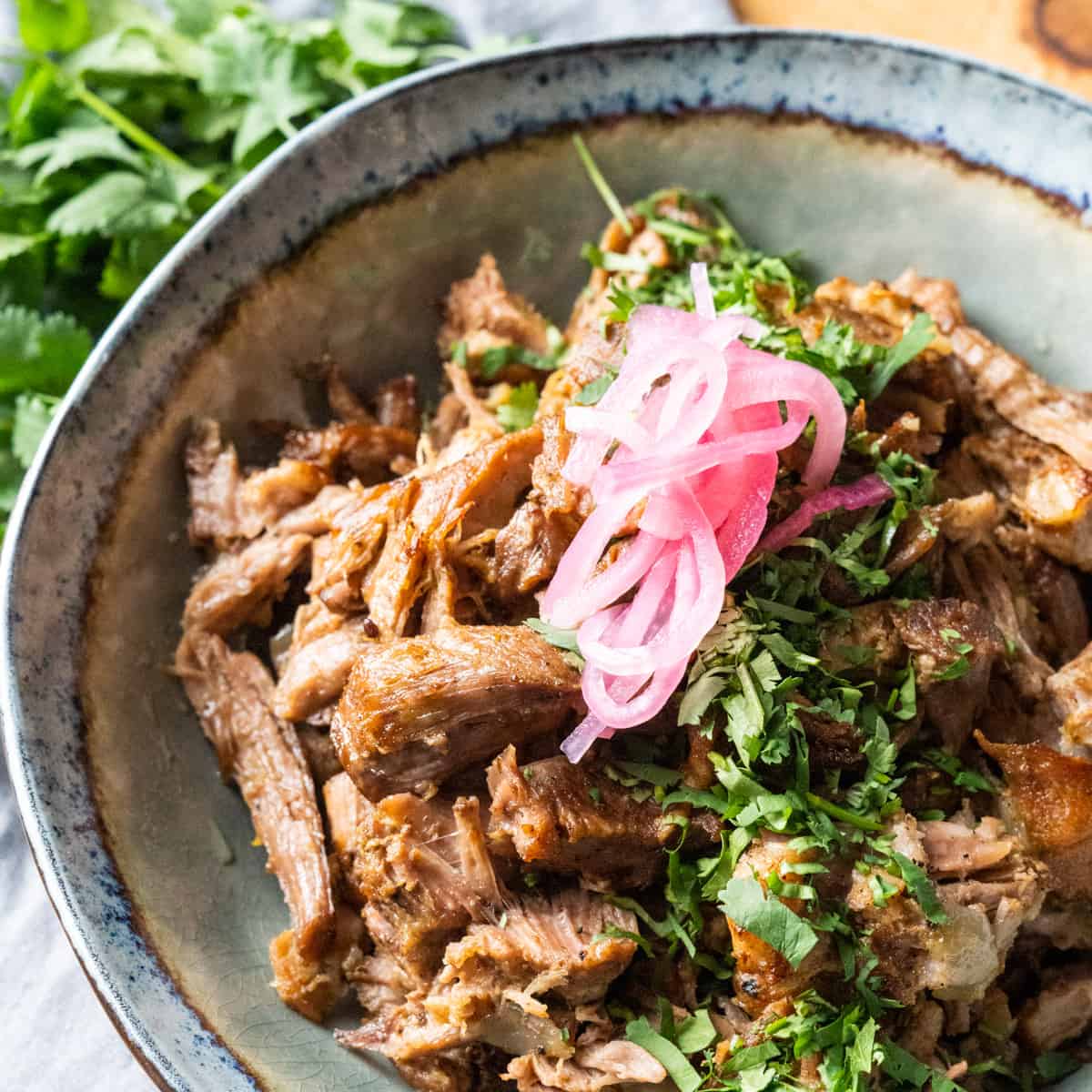 Family style bowl of carnitas garnished with cilantro and pickled red onions