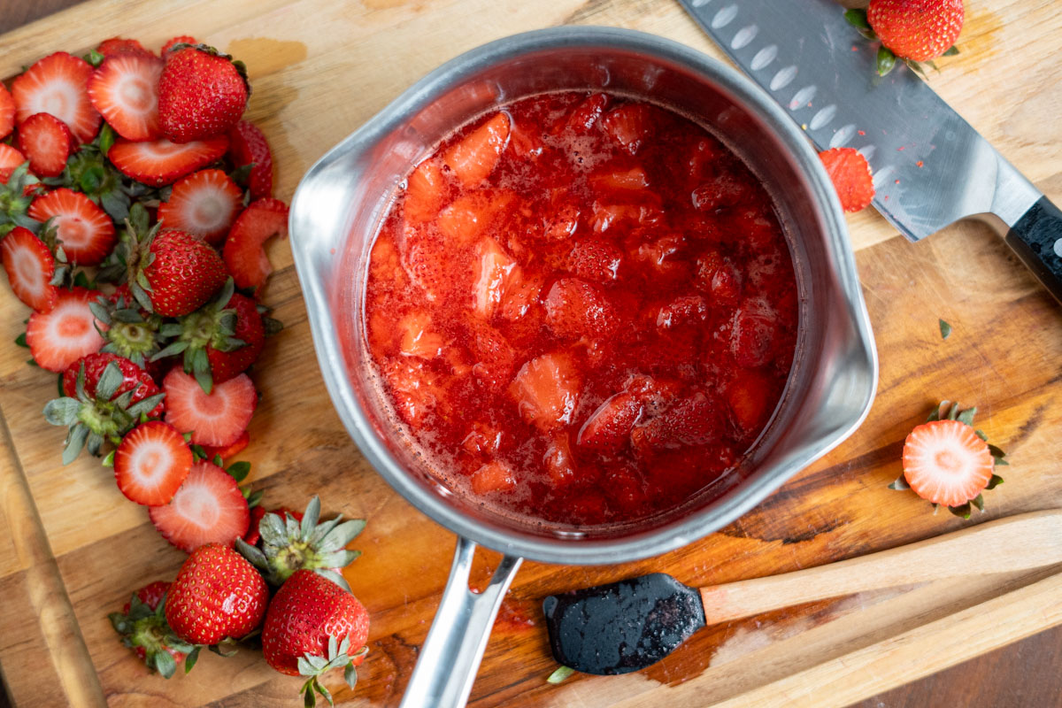 small pot of strawberry compote on a wooden cutting board around sliced berries and a knife