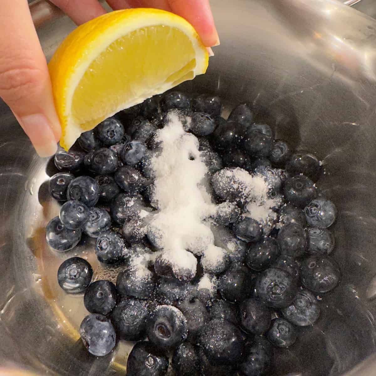 blueberries in a pot with sugar, salt, and a lemon being squeezed over top