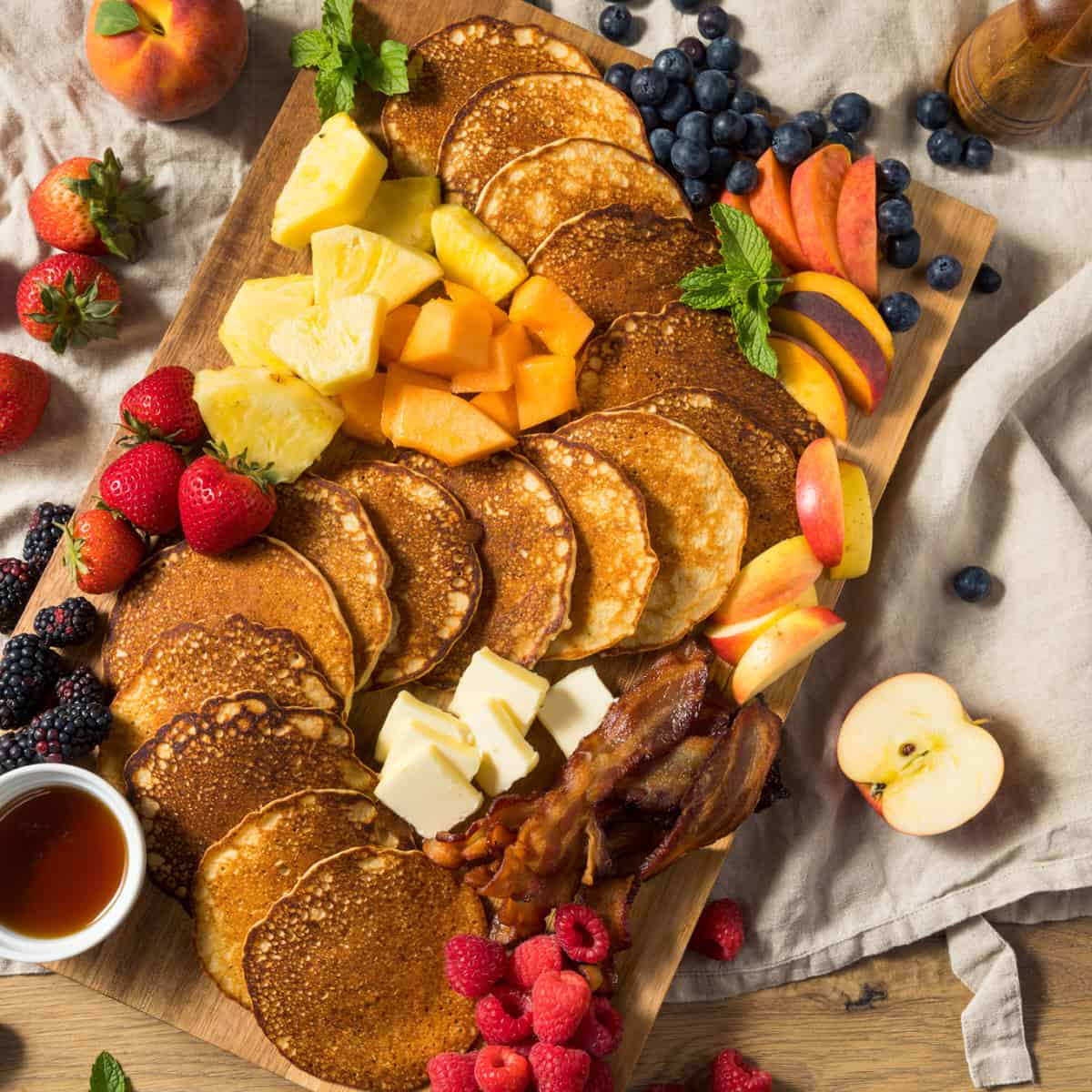 a charcuterie board covered with pancakes, bacon, fresh fruit, and butter