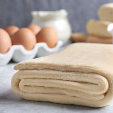 Close-up of folded puff pastry dough on a marble surface with eggs in the background.