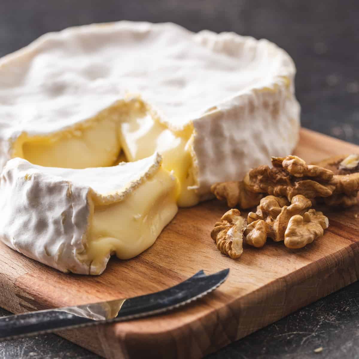 a wheel of brie with a triangular slice removed on a wooden cutting board with walnuts 