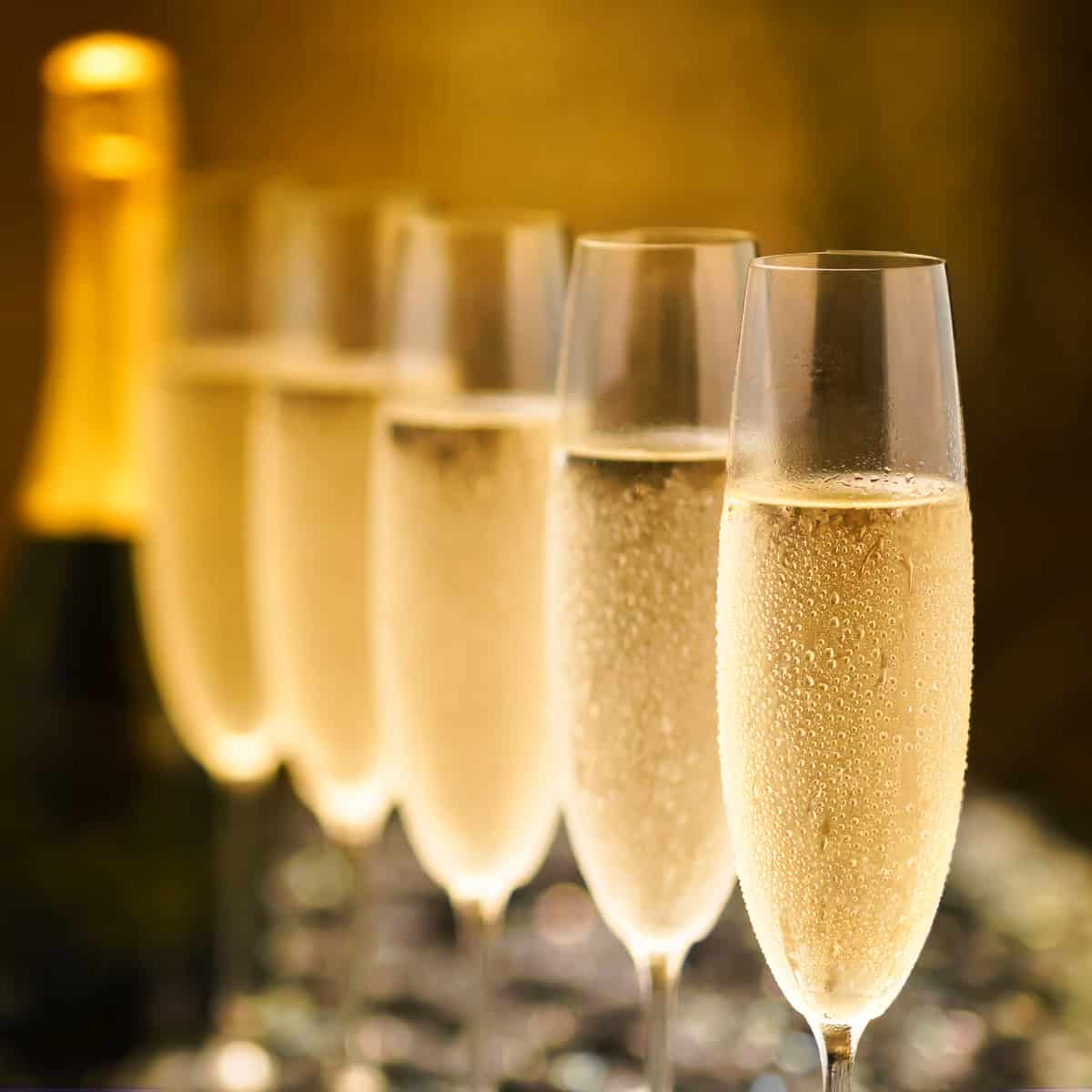 five champagne flutes filled with an unopened bottle of champagne in the background