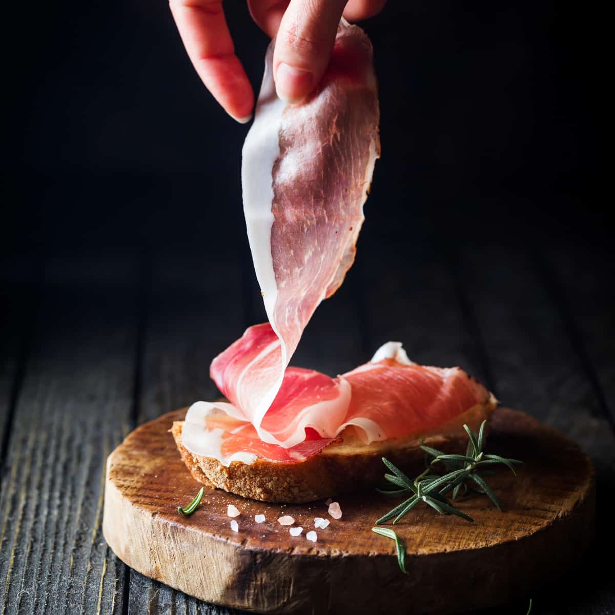 Prosciutto being placed on a wooden circle charcuterie board
