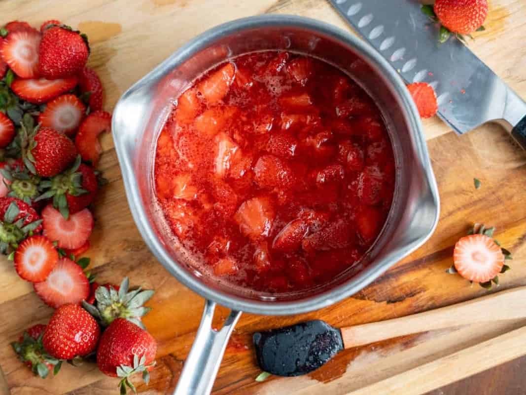 Homemade strawberry compote simmering in a saucepan, surrounded by fresh sliced strawberries on a wooden cutting board.